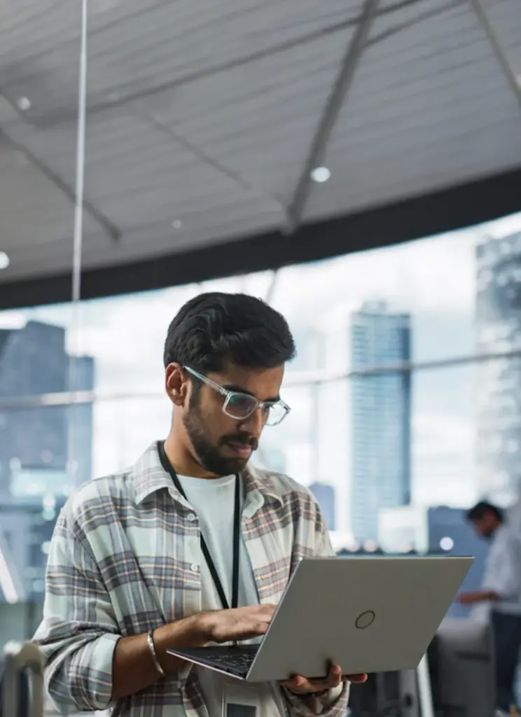 Young professional web developer reviewing website on laptop in modern open office with city skyline – expert website design and development services